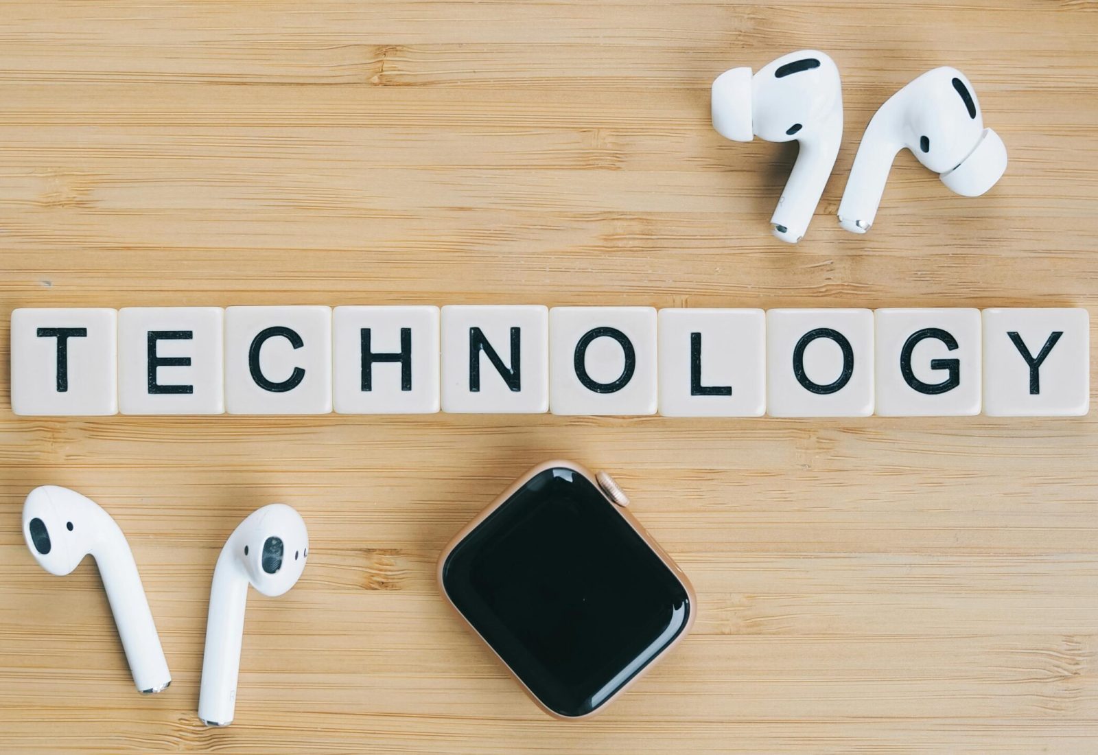 Flat lay of wireless earphones and smartwatch next to TECHNOLOGY tiles on a wooden table.